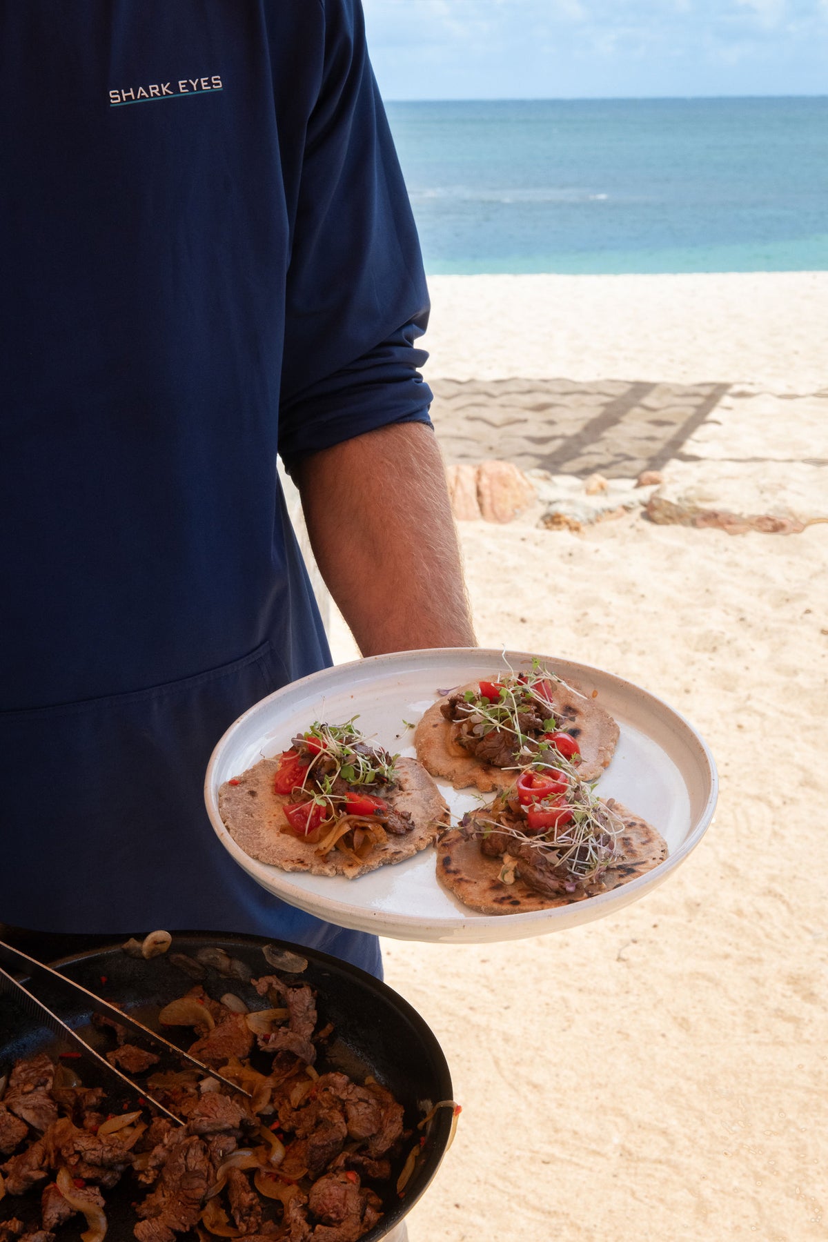 Chef Glen in a Shark Eyes hoodie holding a plate and a pan on the beach